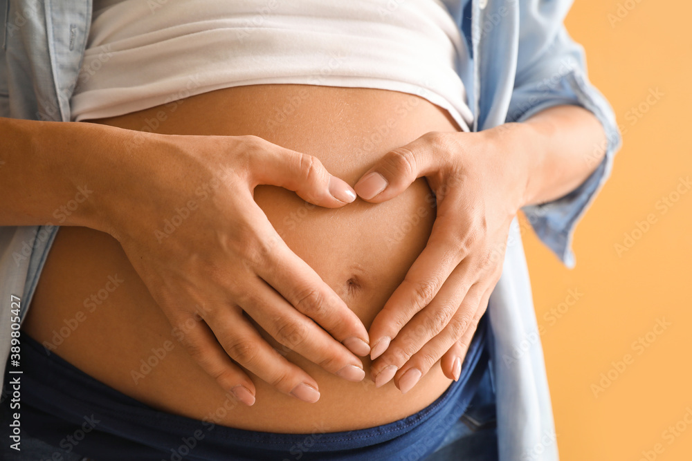 Beautiful pregnant woman making heart with her hands on color background, closeup