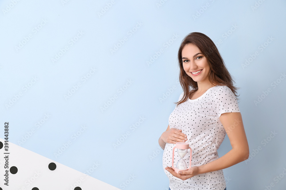 Young pregnant woman with hourglass on color background