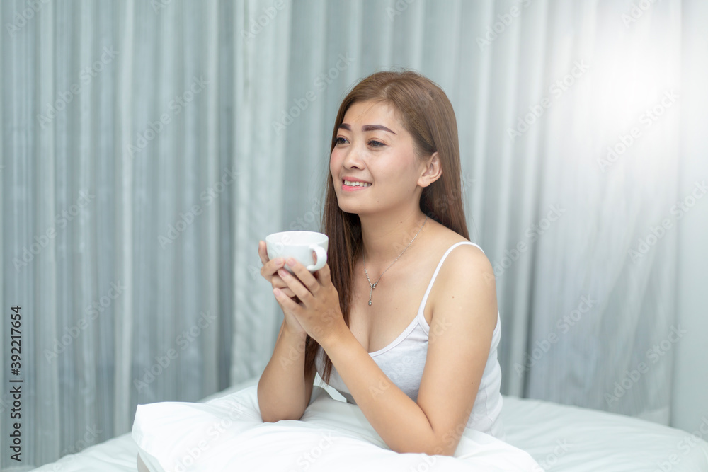 An Asian woman sipping on a morning coffee after waking up in her private bedroom.