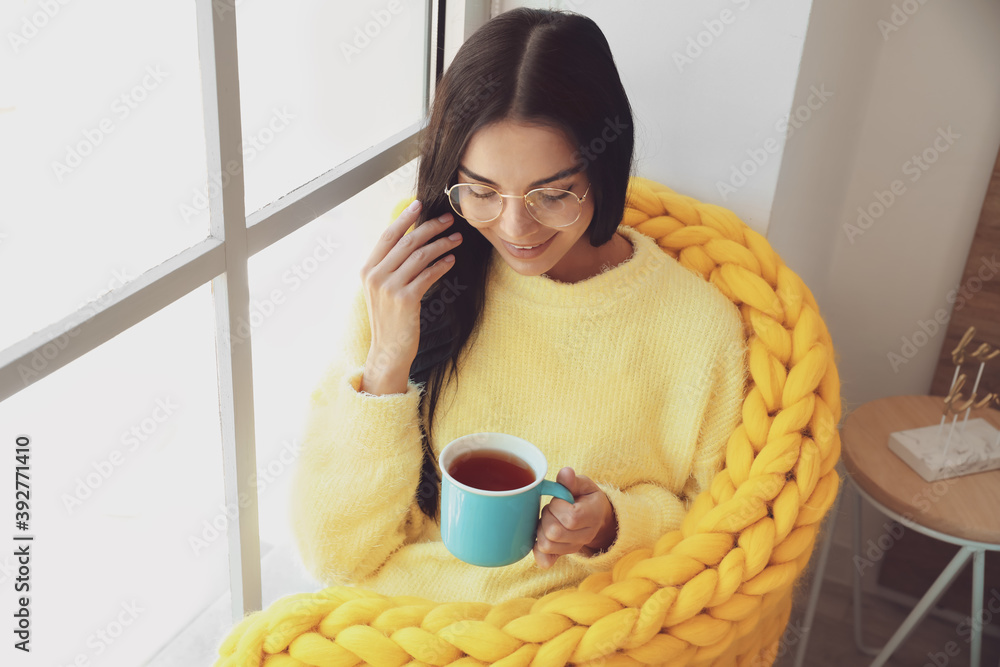 美丽的年轻女子裹着温暖的格子在窗户边喝茶