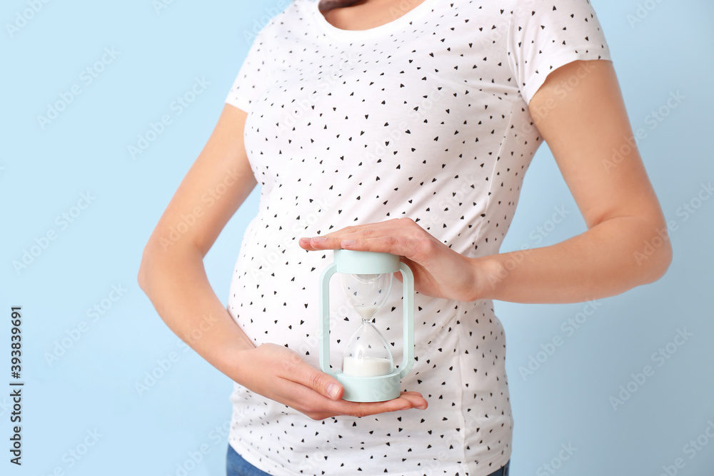 Young pregnant woman with hourglass on color background