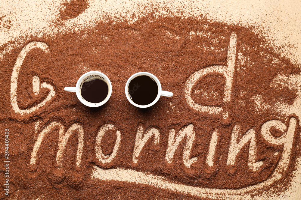 Text GOOD MORNING written on coffee powder and cups of drink on color background