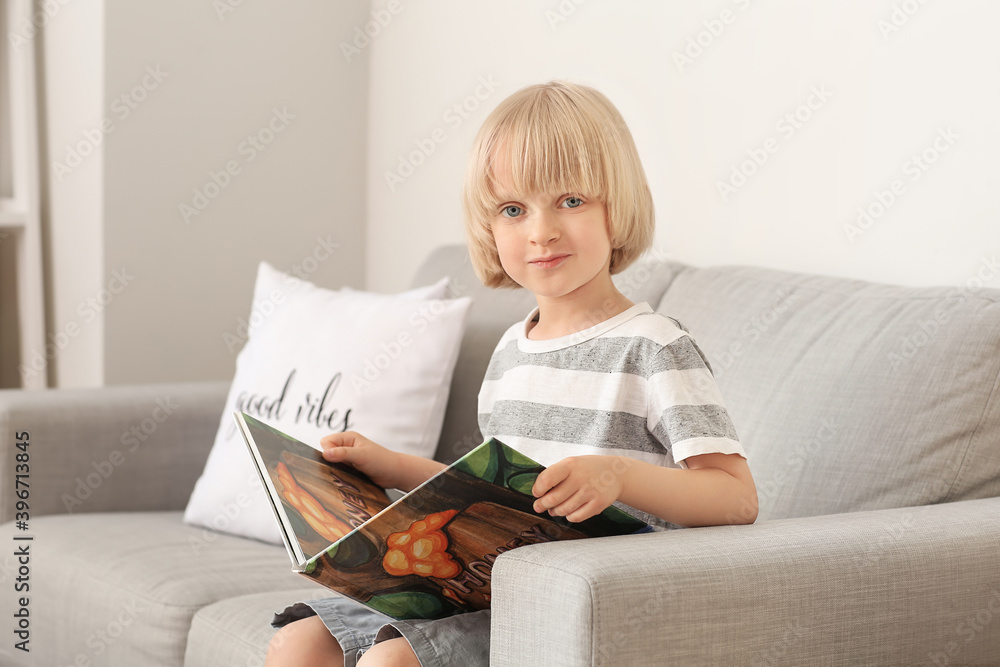 Cute little boy reading book on sofa at home