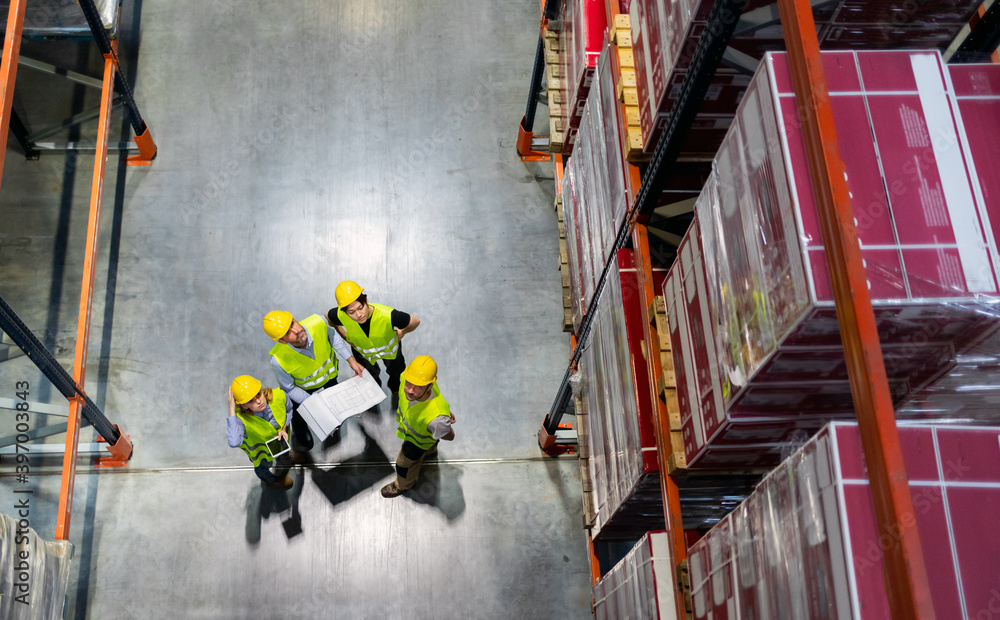 Warehouse manager and workers consult plans between tall shelves, above view