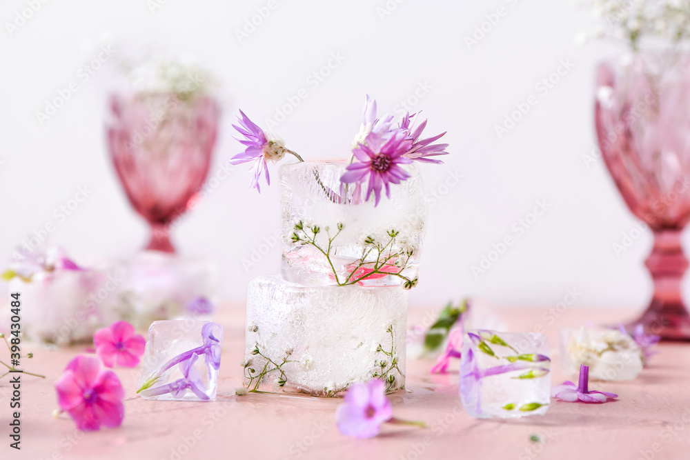 Glasses with beautiful flowers frozen in ice on color background