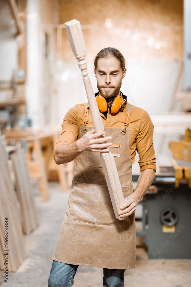 Handsome carpenter in uniform working with wood, checking the quality of the wooden baluster at the 