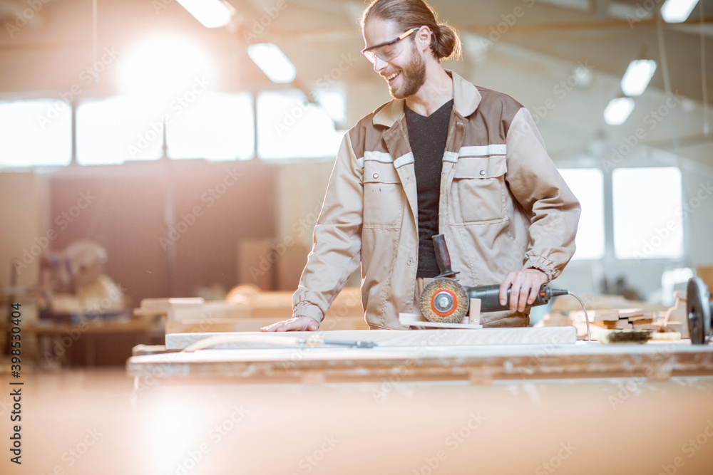 Handsome carpenter in uniform brushing wood with hand machine at the carpentry manufacturing