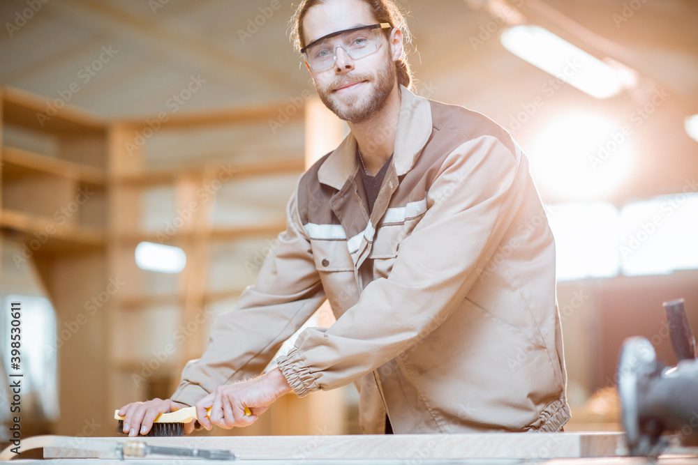 Portrait of a handsome carpenter in uniform brushing wood with hand brush at the carpentry manufactu