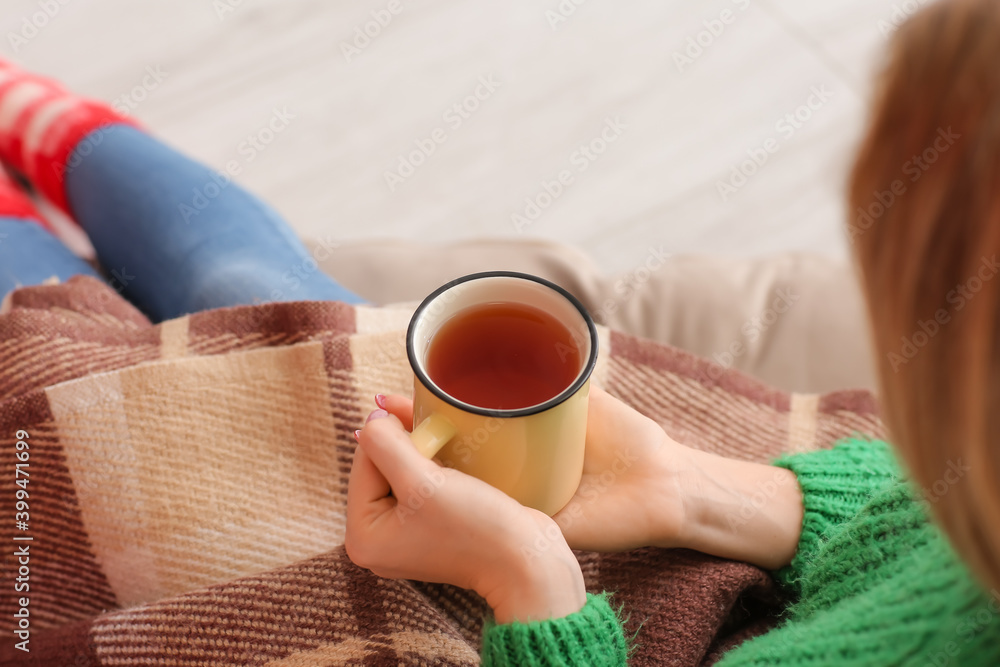 Young woman covered with warm plaid drinking hot tea at home