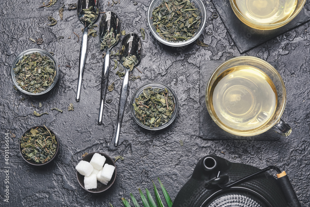 Cups of green tea, spoons and bowls with dry leaves on dark background