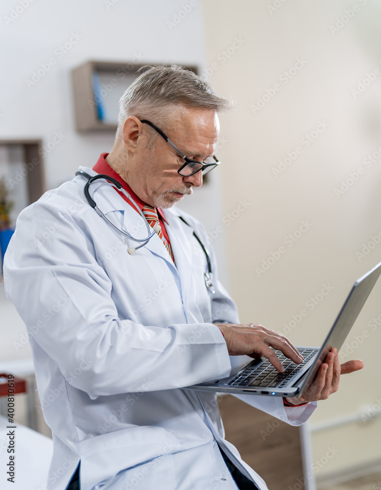 Professional medic types on laptop. Doctor in scrubs and stethoscope on neck works in clinic office.