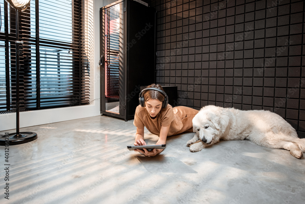 Young relaxed woman works on a digital tablet, lying with a dog on the floor at home music studio. C