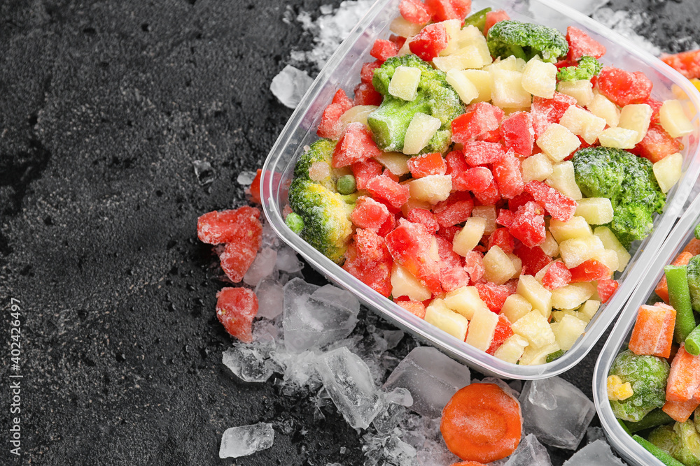 Plastic containers with frozen vegetables and ice on dark background