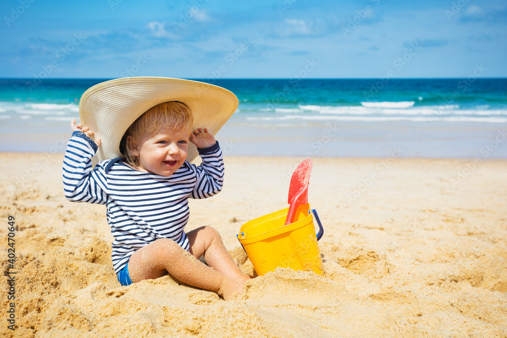 Happy little toddler boy sit on the beach wear big straw hat play with plastic bucket