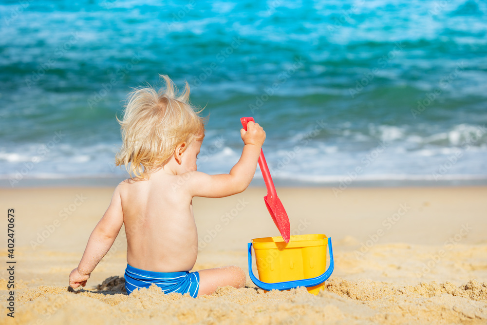 Little happy blond boy play with small bucket hold shovel on the beach near the sea view from back