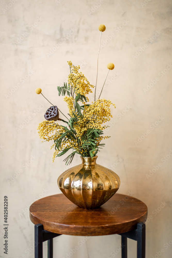 Composition of fresh flowers and herbs in a gold vase on a beige wall background. Bouquet of Acacia,