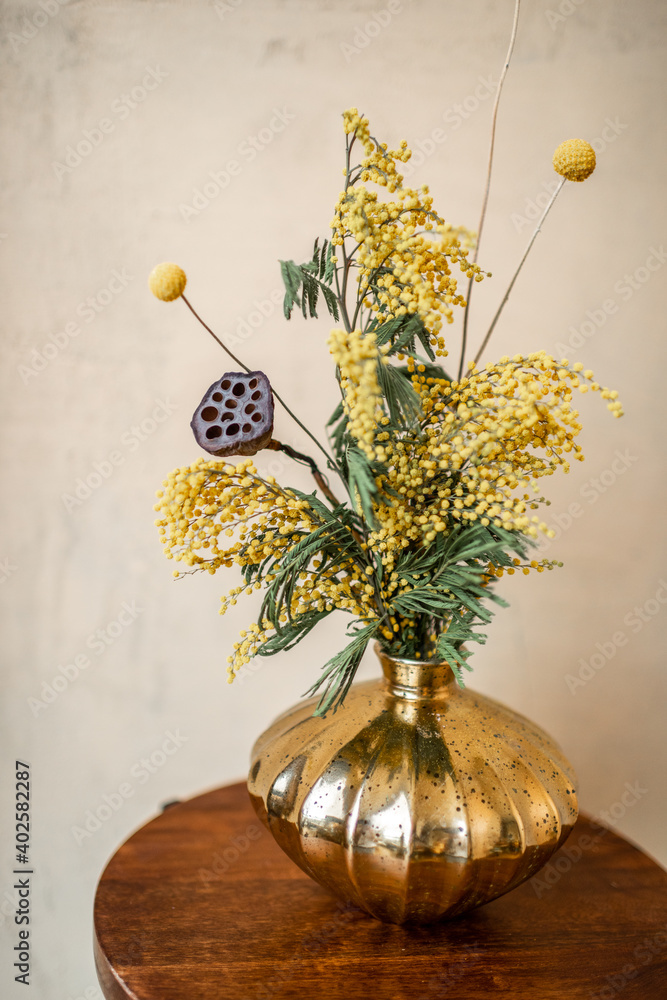 Composition of fresh flowers and herbs in a gold vase on a beige wall background. Bouquet of Acacia,