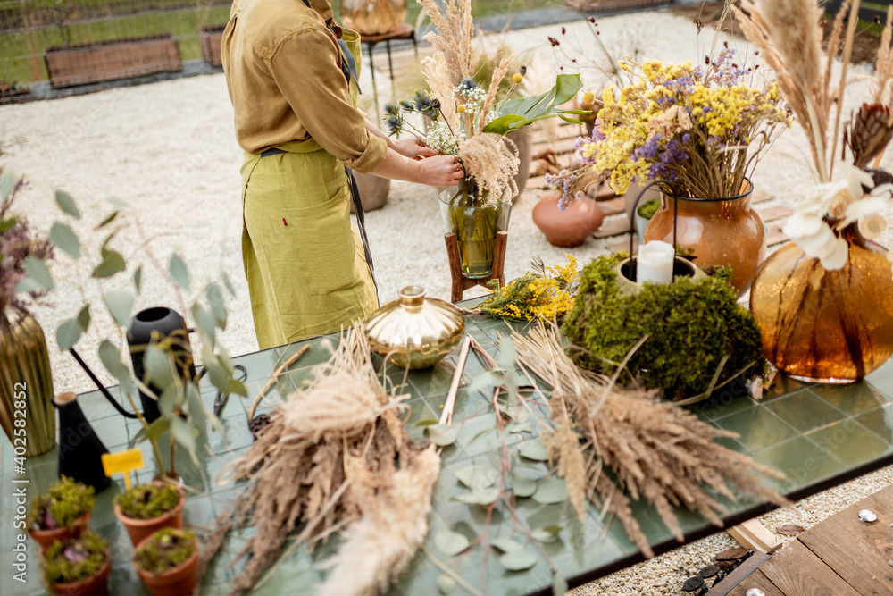 Female making compositions of dried and fresh flowers and herbs on the green table outdoors. Florist