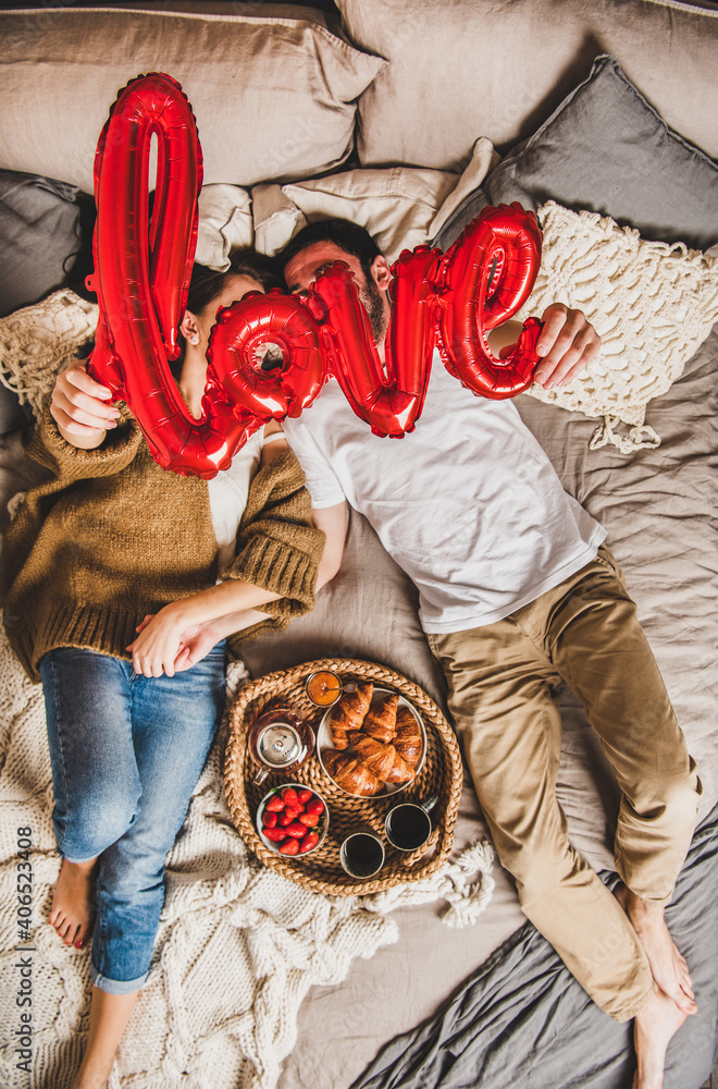 Young happy couple lying on pastel colored blankets barefoot in bed, having breakfast with fresh cro