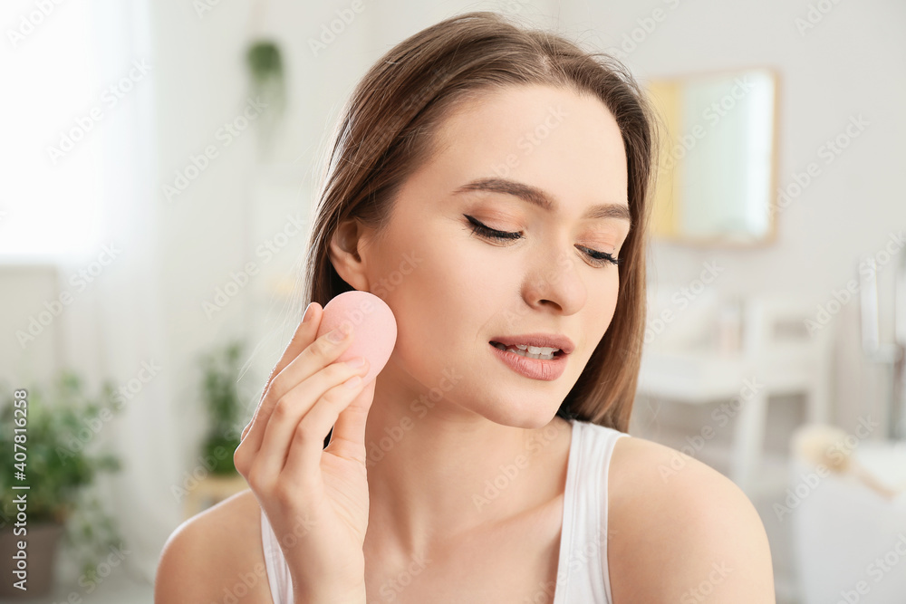 Beautiful young woman with sponge applying makeup at home