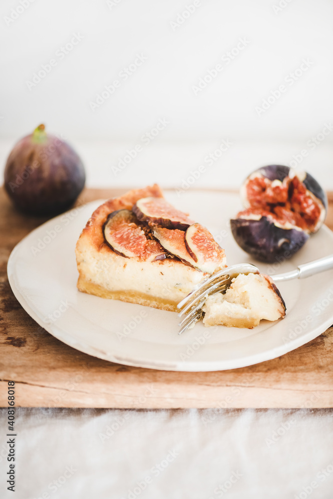 Slice of homemade fig cheesecake with fresh figs on white plate over rustic wooden board, white wall