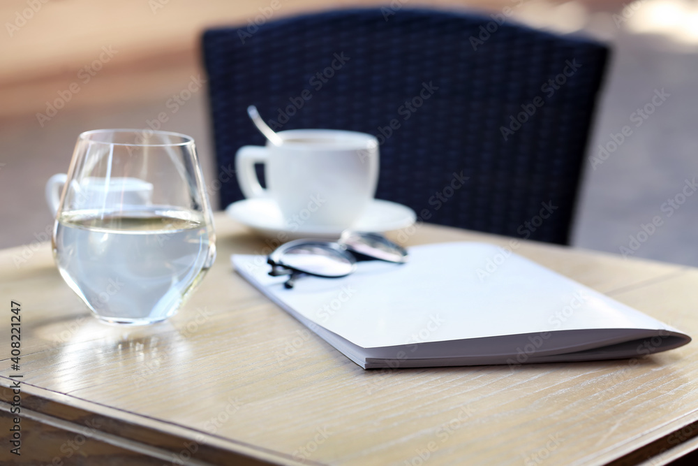 Blank magazine, glass of water and cup with coffee on table in cafe outdoors