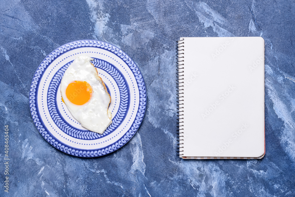 Plate with tasty fried egg and blank notebook on color background