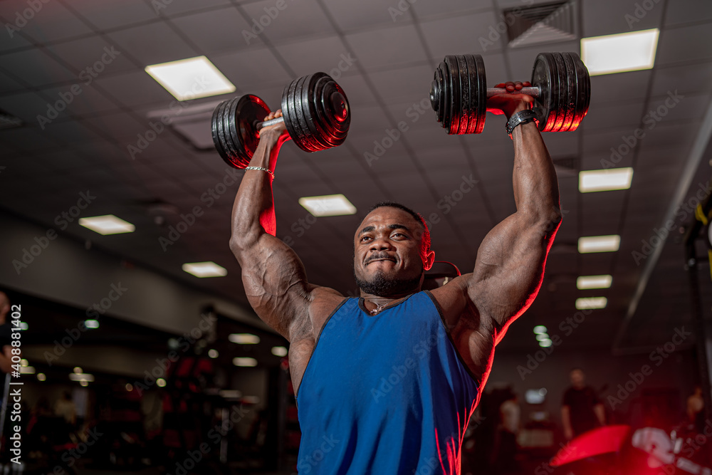 African american athlete pumping dumbbels over head. Selective focus from below on strong bodybuilde