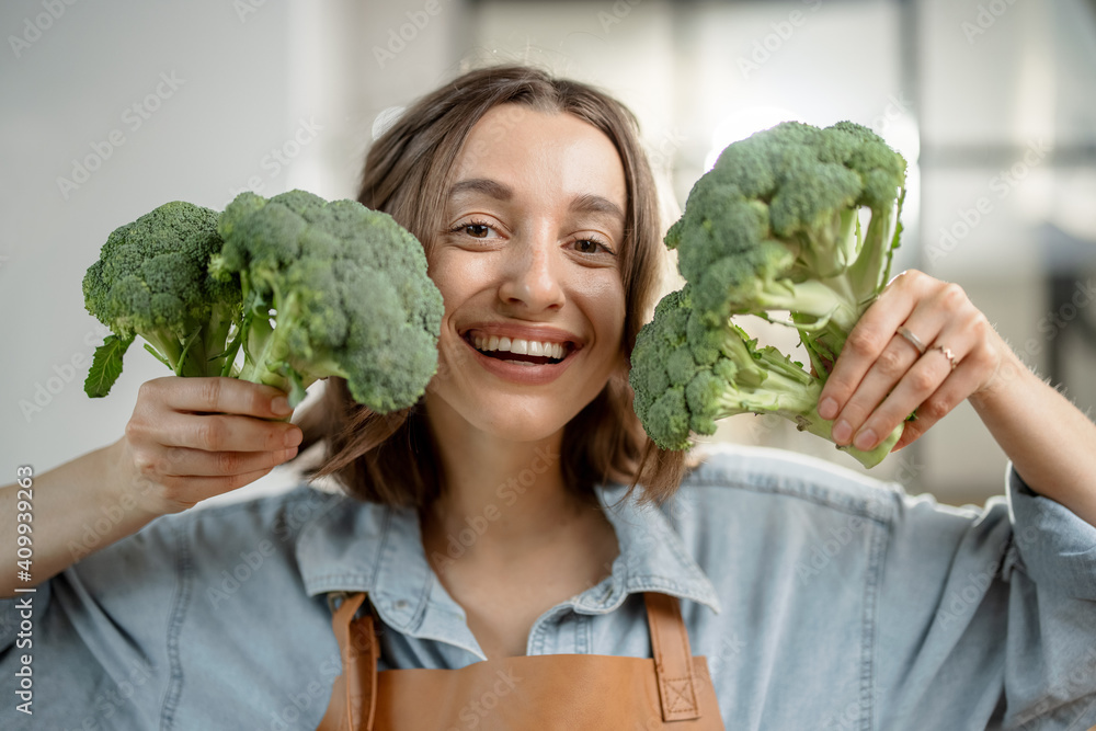 Portrait of pretty smiling woman in apron with fresh broccoli on the kitchen. Healthy cooking concep