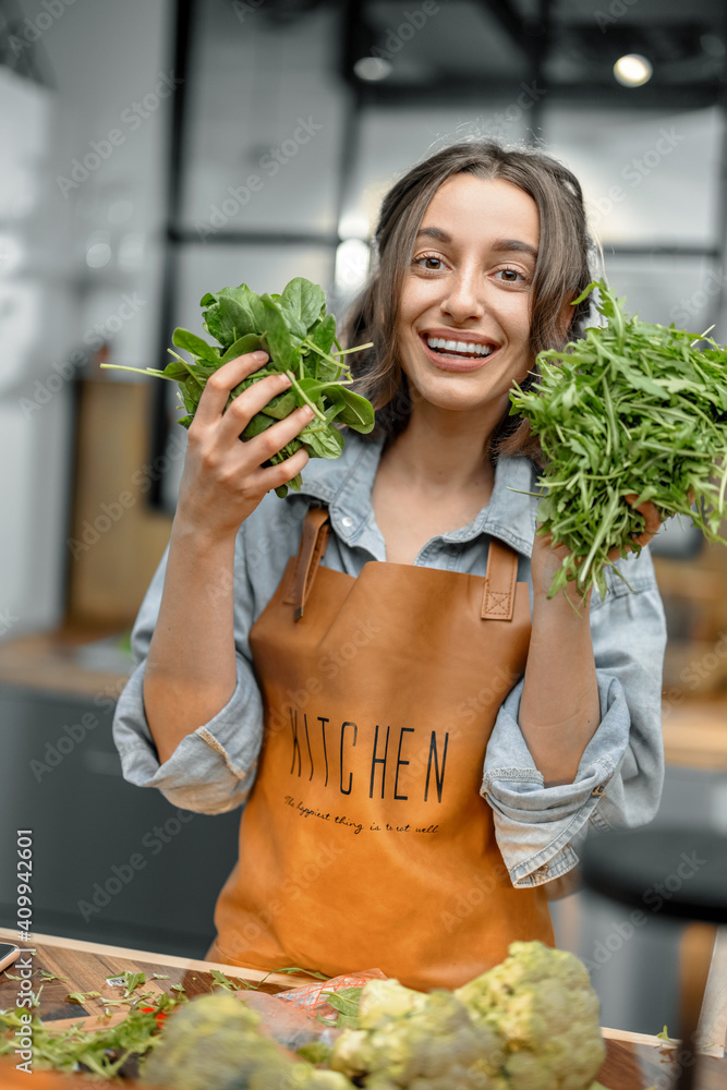 Portrait of cheerful woman in apron with fresh spinach and arugula on the kitchen. Healthy cooking c