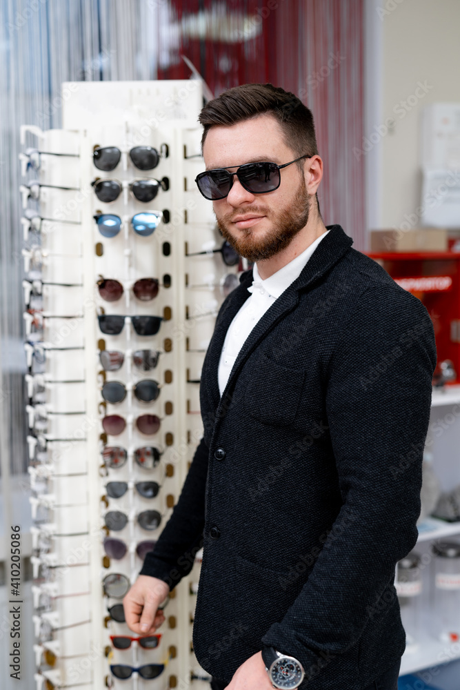 Handsome man model chooses fashion sunglasses in optics. Wide assortment of modern eyewear in shop.