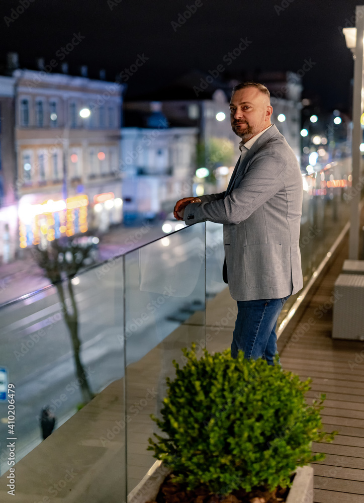 Senior elegant handsome man stands on modern balcony with glass handrail. Evening city blurred backg