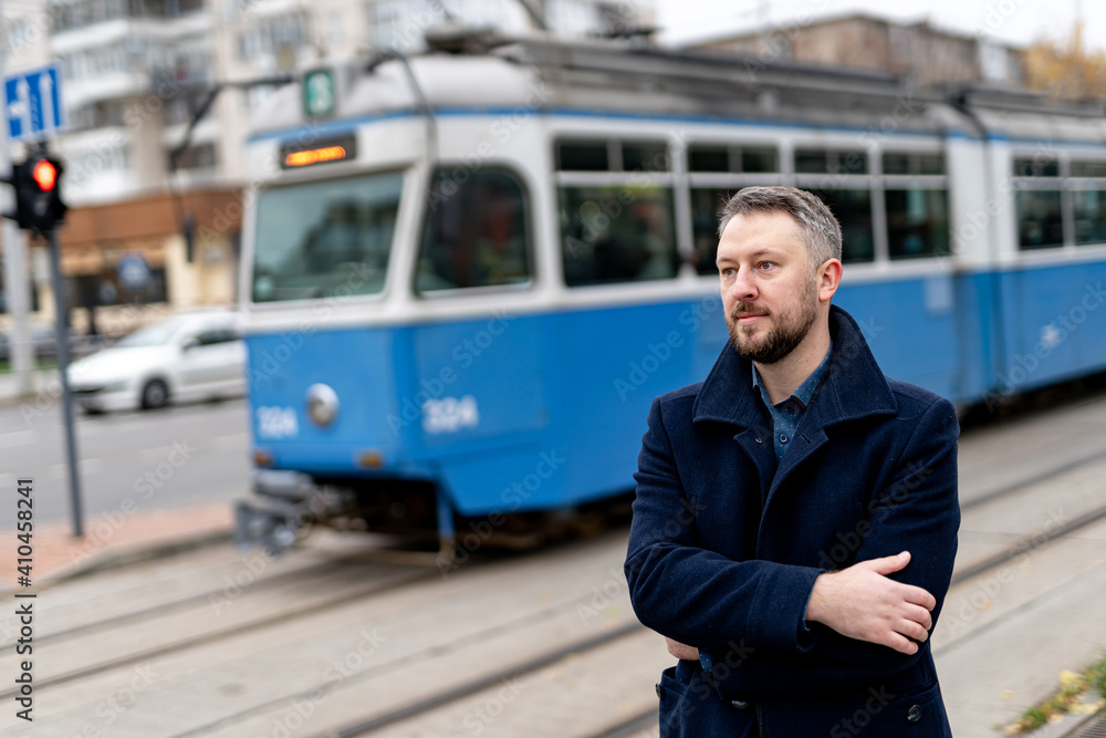 Handsome male in coat stands on the city blue tram background. Man waits for green light to cross th