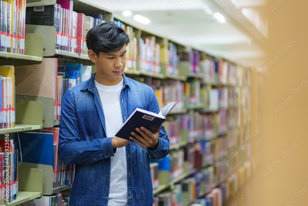 Portrait of Smart Asian man university student reading book between bookshelves in campus library wi