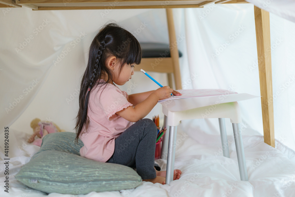 Asian cute little girl drawing in paper while lying in a blanket fort in living room at home for per