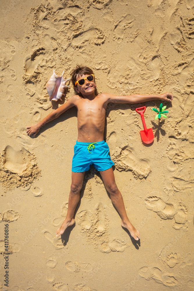 Boy lay on the sand with shell and plastic scoop wearing shades smiling, view from above