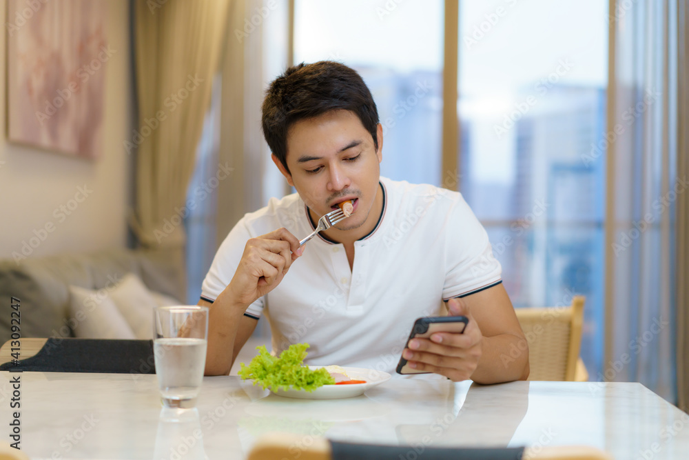 一名亚裔男子坐在椅子上吃美国早餐，打电话，查看电子邮件