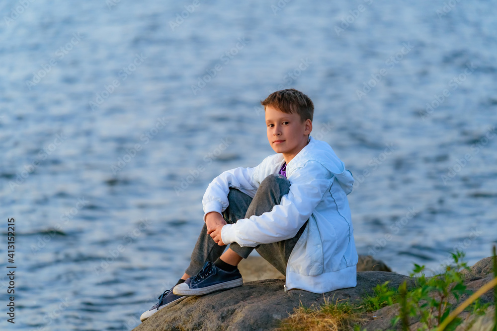 Serious boy in a white jacket with a hood and jeans is sitting on a rock near the river. Closeup.