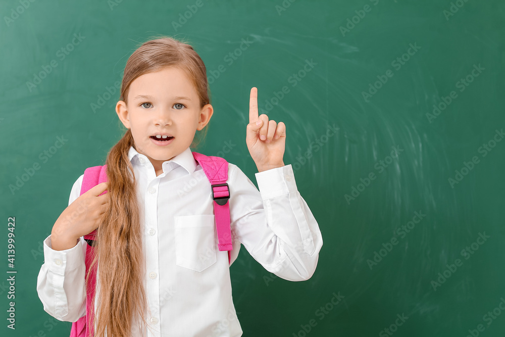 Little schoolgirl with raised index finger near chalkboard in classroom