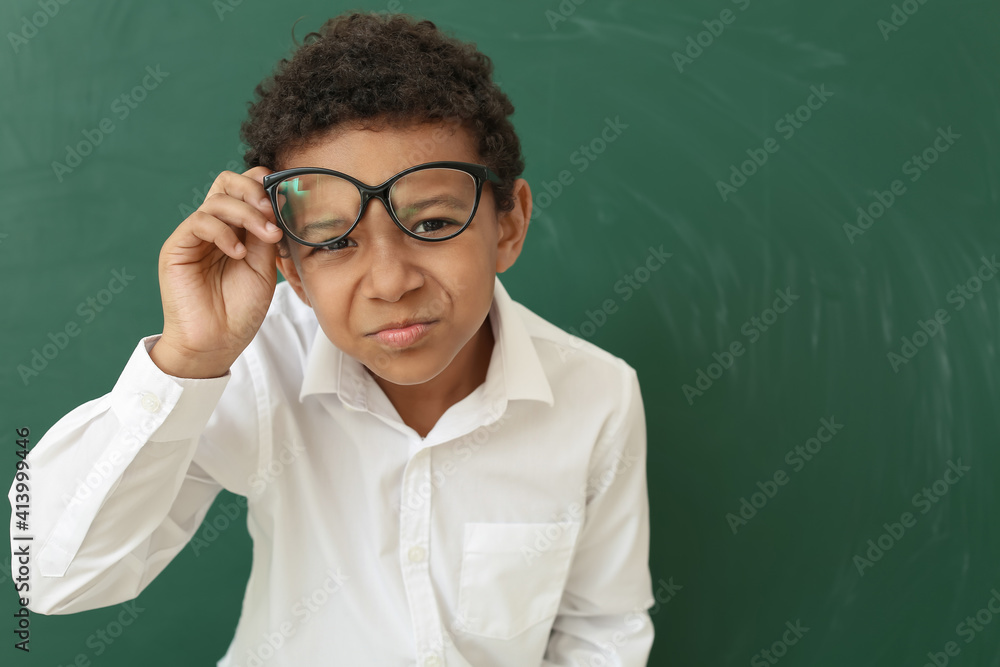 Curious little African-American schoolboy with eyeglasses near chalkboard in classroom