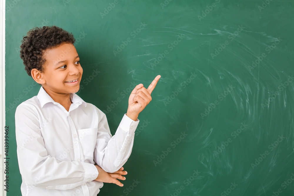 Little African-American schoolboy showing something near chalkboard in classroom