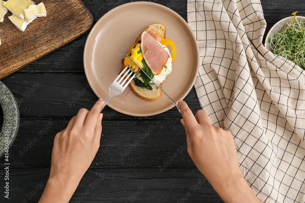 Woman eating tasty sandwich with florentine egg and bacon on dark wooden background