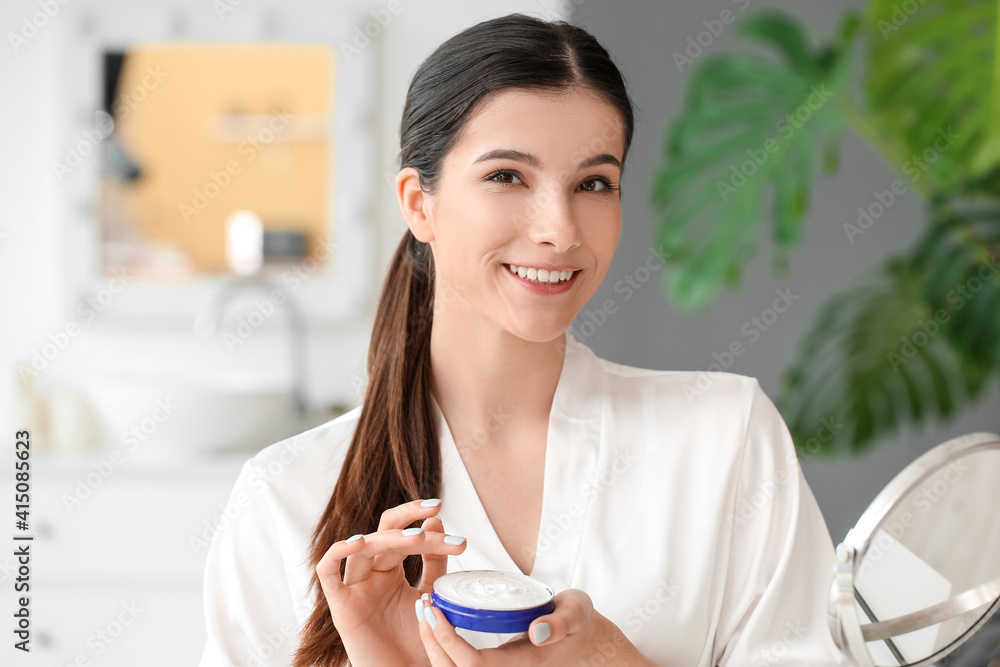 Beautiful young woman applying facial cream in bathroom