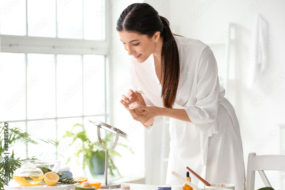 Beautiful young woman applying facial cream in bathroom