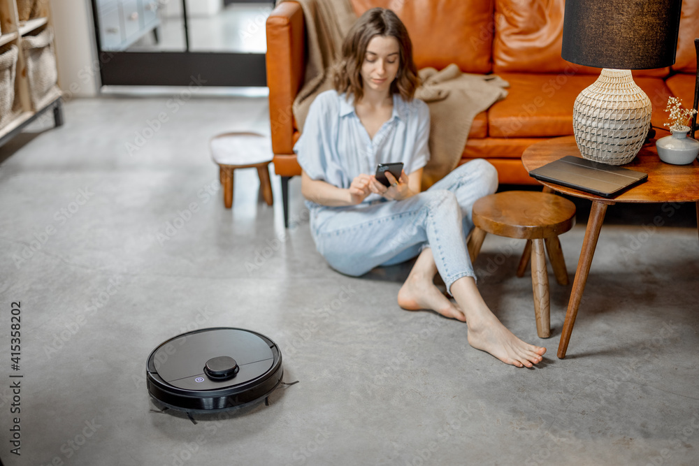 Black robotic vacuum cleaner cleaning the floor while woman sitting near sofa and using phone. Smart