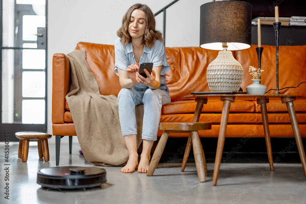 Black robotic vacuum cleaner cleaning the floor while woman sitting on sofa and looking on her house