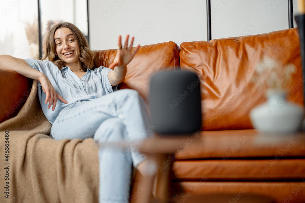 Happy pretty woman speaking with black audio assistant column on the coffee table while sitting on f