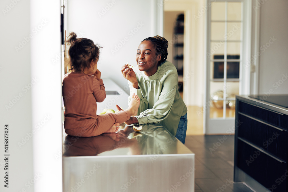 Smiling mom and her little daughter eating fruit at home