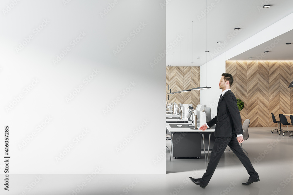 Businessman in black suit goes by modern workplaces in sunny open space office with blank light wall