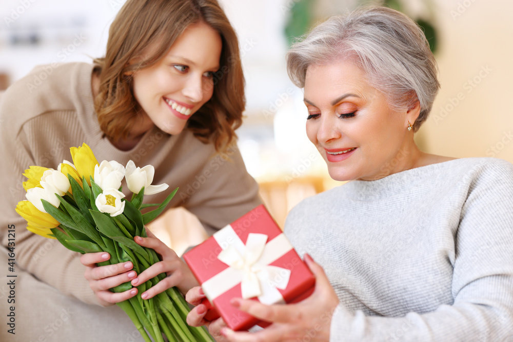 Young woman giving flowers and gift to senior mother